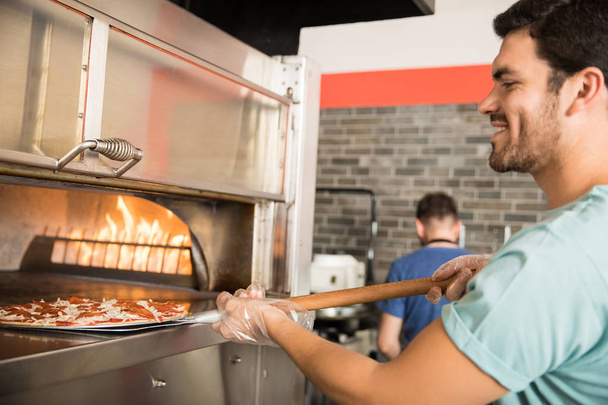 Szczegół portret wesoły kucharz na sobie rękawice, wprowadzenie surowego przygotowane pizzy w piecu przy użyciu skórki w sklepie pizza - Zdjęcie, obraz