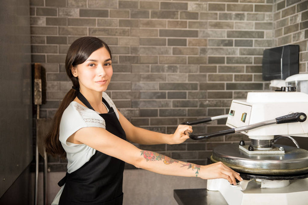 Beautiful chef woman wearing black apron using dough press machine to make flat pizza bread in kitchen at pizza store while looking at camera - Valokuva, kuva