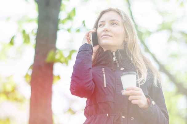 Hablando por teléfono, Una chica está caminando por la mañana en un parque con un teléfono inteligente y café, tonificación ligera
 - Foto, imagen