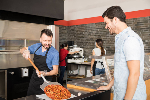 Bonito homem sorrindo olhando para a ordem de pizza recém-assado que está sendo colocado em servir prato pelo chef usando uma casca no balcão da cozinha do restaurante
 - Foto, Imagem