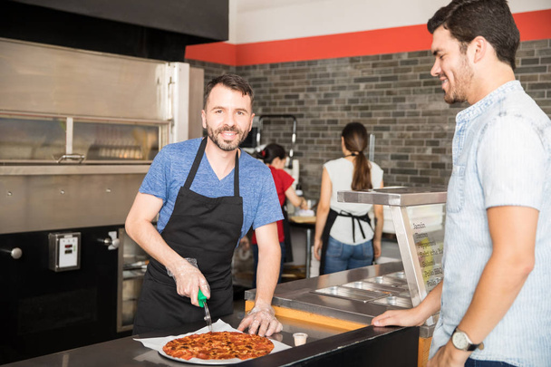 Feliz chef cortando pizza en rodajas mirando a la cámara mientras el guapo cliente espera pizza en la pizzería
 - Foto, imagen