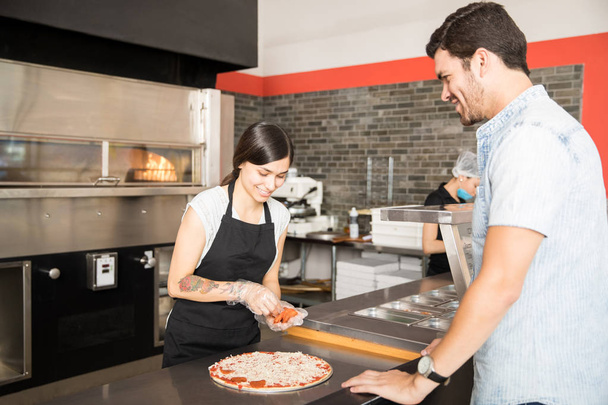 Beautiful chef preparing pepperoni pizza for customer standing at kitchen counter in pizza shop - Photo, Image