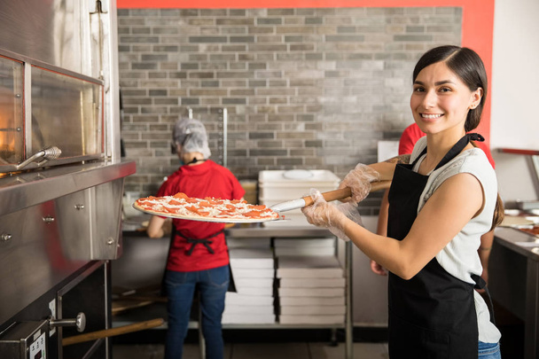 Smiling chef wearing uniform putting raw pizza in modern oven for baking while looking at camera and staff working in background in pizza shop - Foto, Bild