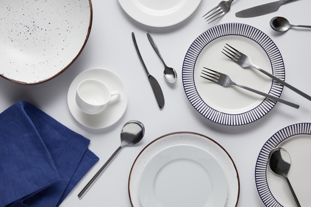 top view of different ceramic plates, cup, kitchen towel, forks, spoons and knife on white table  - 写真・画像