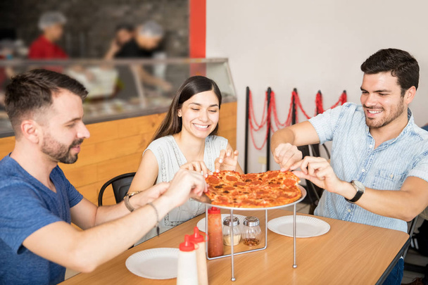 Two handsome men with cheerful beautiful woman enjoying pizza party at restaurant while picking up slices - Photo, Image