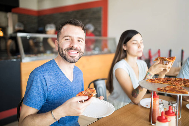 Portrait of young handsome man holding pepperoni pizza slice in hand sitting in restaurant with friends in background while looking at camera - Zdjęcie, obraz