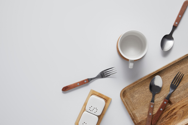 top view of cup, spoons, forks, wooden tray, saltcellar and pepper caster on white table  - Foto, immagini