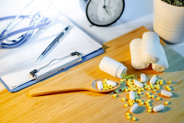 Таблетки, выливающиеся из пузырька с марлевым бинтом, фармацевтическая деревянная ложка, стетоскоп, планшет и ручка на столе врача. Лекарство по рецепту или медицинская диагностика и лечение
 - Фото, изображение