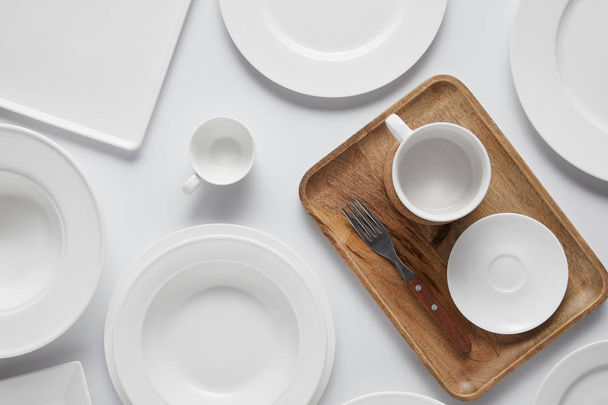 top view of cup, different plates, wooden tray, bowl and fork on white table - Fotoğraf, Görsel