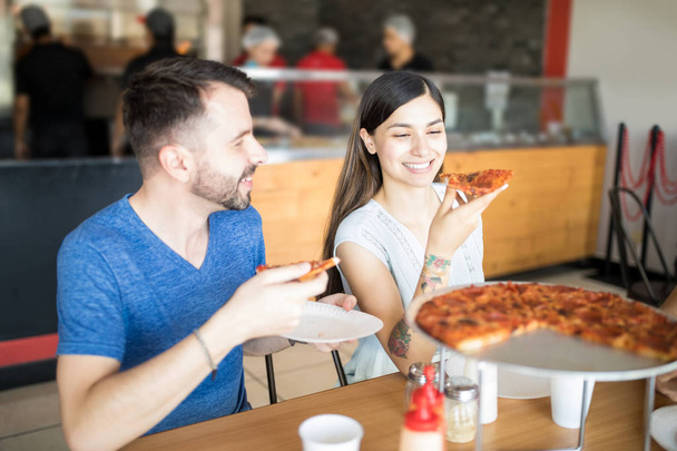 Mujer atractiva y hombre guapo disfrutando de pizza de pepperoni en la pizzería durante el almuerzo mientras sostiene rebanadas de pizza y habla
 - Foto, Imagen
