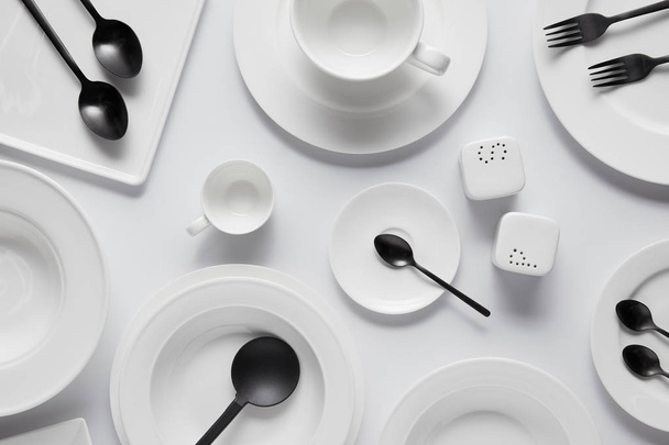 vista superior de cucharas negras, tenedores, cuchara de ensalada, diferentes platos, tazón, bodega de sal, echador de pimienta y taza en mesa blanca
 - Foto, Imagen