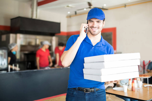 Lächelnder, gutaussehender junger Mann in blauem Hemd und Mütze, der im Restaurant vier Pizzaschachteln in der Hand hält und telefonisch bestellt - Foto, Bild