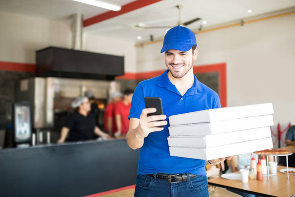 Красивый веселый мужчина проверяет адрес доставки, держа в руках стопку коробок с пиццей для доставки в ресторан
 - Фото, изображение