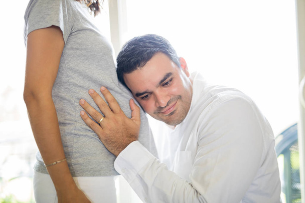 Homem sorridente ouvindo a barriga da mulher grávida em casa
 - Foto, Imagem
