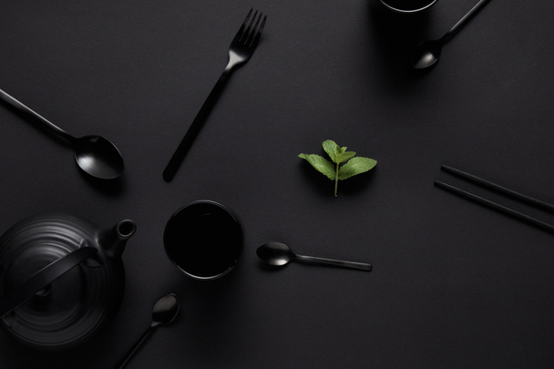 Top näkymä musta teekannu, syömäpuikot, eri lusikat, haarukka, kuppi ja mintunlehtiä musta pöytä
 - Valokuva, kuva