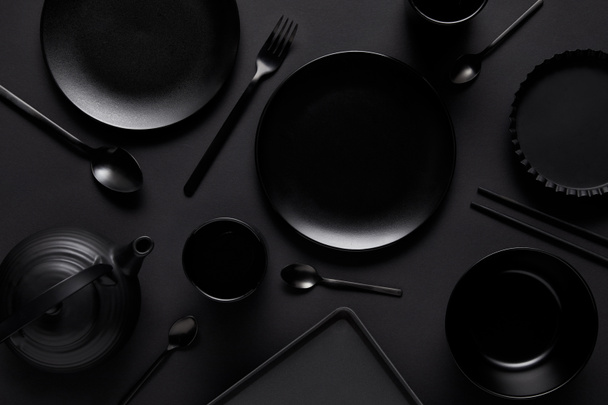 top view of black teapot, plates, bowl, cups, tray, spoons, chopsticks, fork and baking dish on black table  - Foto, Bild