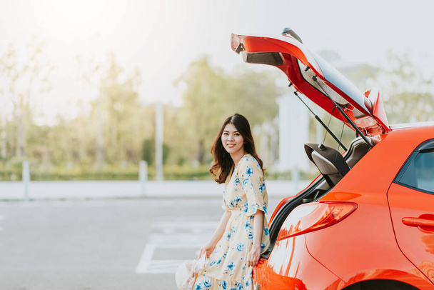 Счастливая азиатка-путешественница сидит на багажнике автомобиля во время отпуска
 - Фото, изображение