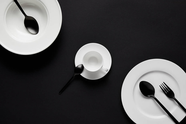 top view of various plates, cup, black spoons and fork on black table - Фото, зображення