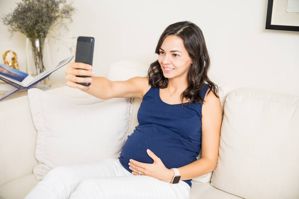 Беременная женщина делает селфи на смартфоне, сидя дома на диване
 - Фото, изображение