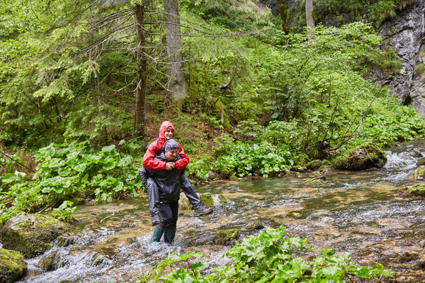 Mies korkeissa saappaissa reppuselässä kantamassa poikaa joen yli
 - Valokuva, kuva