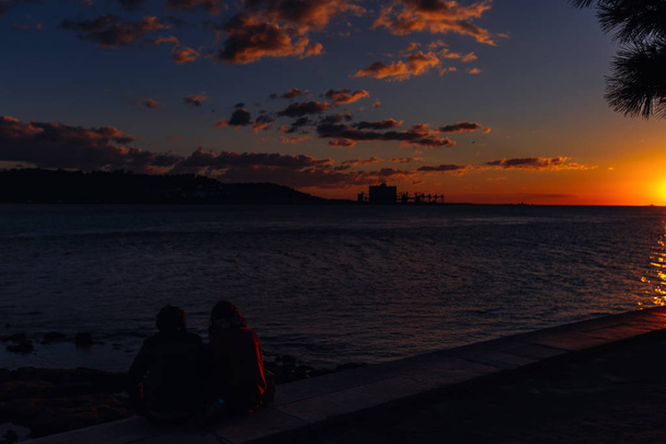 dwóch ludzi siedzi na brzegu rzeki podziwiając piękny zachód słońca. odcienie niebieskie i pomarańczowe niebo i sylwetki przeciwległym brzegu - Zdjęcie, obraz