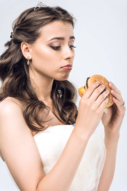 sad young bride in wedding dress looking at burger in hands isolated on white - Fotografie, Obrázek