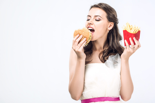 hangry young bride in wedding dress eating burger and french fries isolated on white - Foto, imagen