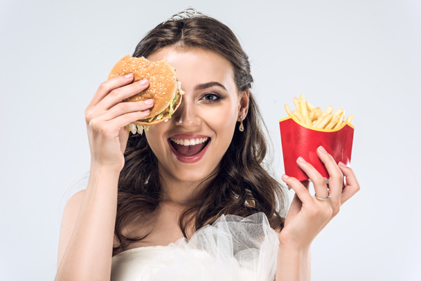 happy young bride in wedding dress with burger and french fries isolated on white - Фото, изображение