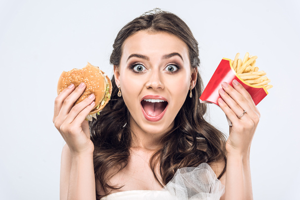 close-up portrait of shocked young bride in wedding dress with burger and french fries looking at camera isolated on white - Foto, immagini