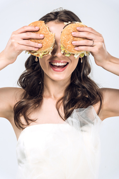 laughing young bride in wedding dress covering eyes with burgers isolated on white - Fotó, kép