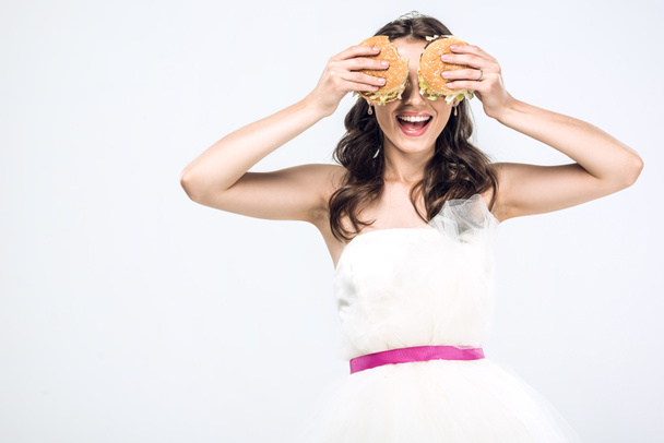 smiling young bride in wedding dress covering eyes with burgers isolated on white - Fotografie, Obrázek