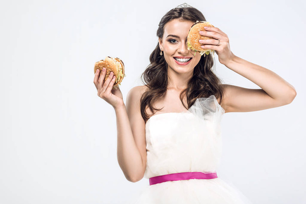 smiling young bride in wedding dress holding burgers in hands isolated on white - Photo, Image