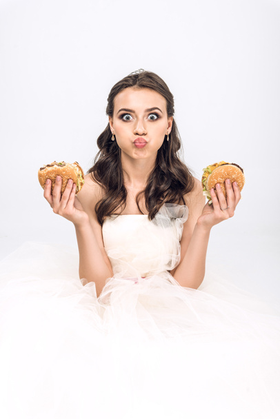 attractive young bride in wedding dress holding burgers in hands while grimacing and looking at camera isolated on white - Foto, afbeelding