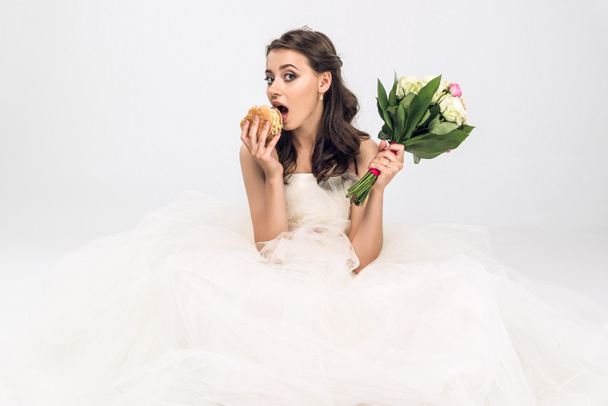 shocked young bride in wedding dress sitting on floor with burger and bridal bouquet in hands on white - Фото, зображення