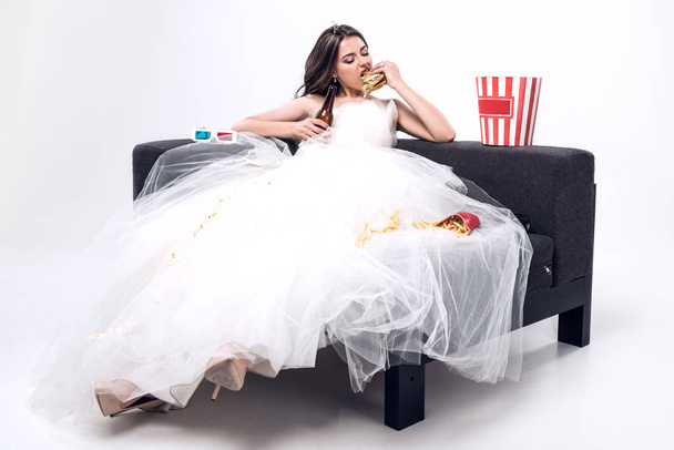 depressed hungry young bride in wedding dress sitting on couch and eating burger with beer on white - Foto, Bild