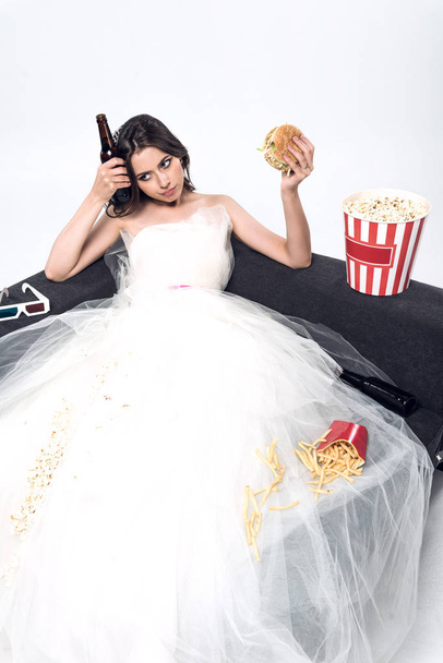 depressive junge Braut im Hochzeitskleid sitzt auf Couch mit Bier und Junk Food auf weiß - Foto, Bild
