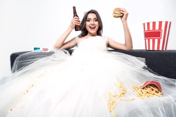 happy young bride in wedding dress sitting on couch with beer and junk food isolated on white - Foto, afbeelding