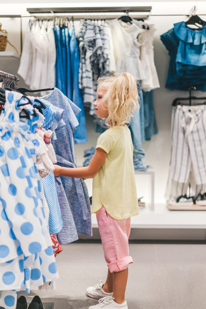 vista lateral de adorable niño pequeño elegir la ropa en la tienda
  - Foto, Imagen