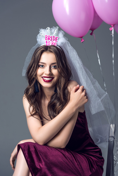 happy future bride in veil for bachelorette party sitting on chair with tied pink balloons isolated on grey - Foto, immagini
