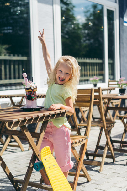 selective focus of smiling child doing peace gesture at table with delicious dessert in cafe - Photo, Image