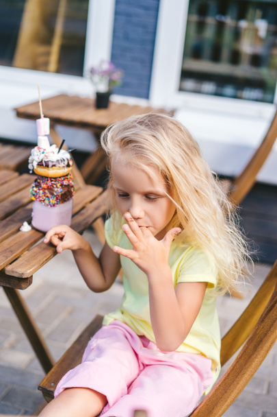 selective focus of little child licking fingers while sitting at table with delicious dessert in cafe - Zdjęcie, obraz