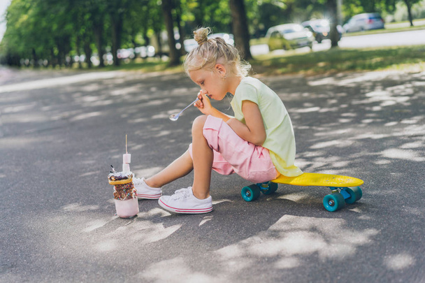side view of little child sitting on skateboard and eating dessert by spoon at street - Valokuva, kuva