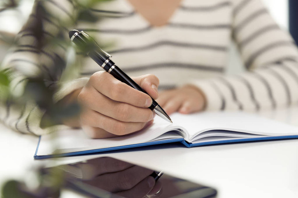 Νεαρή γυναίκα κάθεται στο τραπέζι και γράφοντας στο Σημειωματάριο. Στο τραπέζι είναι smartphone και στο tablet σας. Ελεύθερος επαγγελματίας εργάζεται, γράφει κάτω από νέες ιδέες - Φωτογραφία, εικόνα