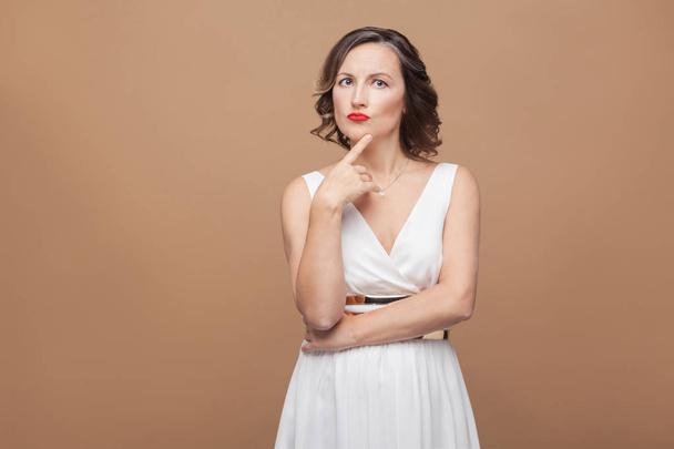 puzzled beautiful middle aged woman in white dress with red lips and dark curly hairstyle  touching her chin and standing against - Zdjęcie, obraz