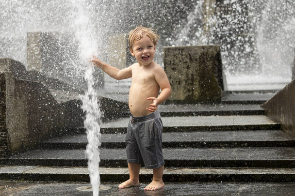 Funny baby boy trying to cauch water stream in fountain. Cute toddler playing in the city fountain. - Photo, Image