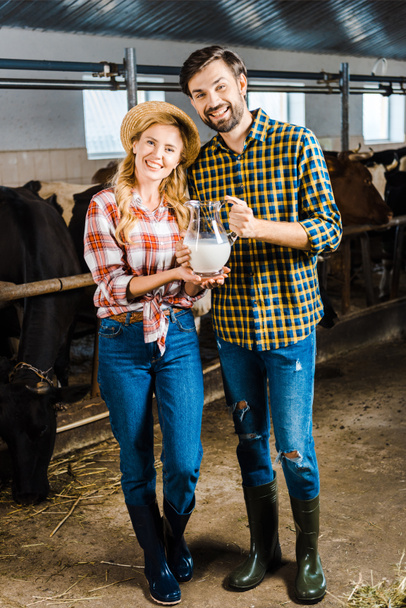 casal sorridente de agricultores segurando jarro de leite em estábulo
  - Foto, Imagem
