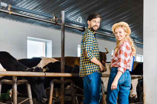 happy couple of farmers looking at camera in stable with cows - Photo, Image