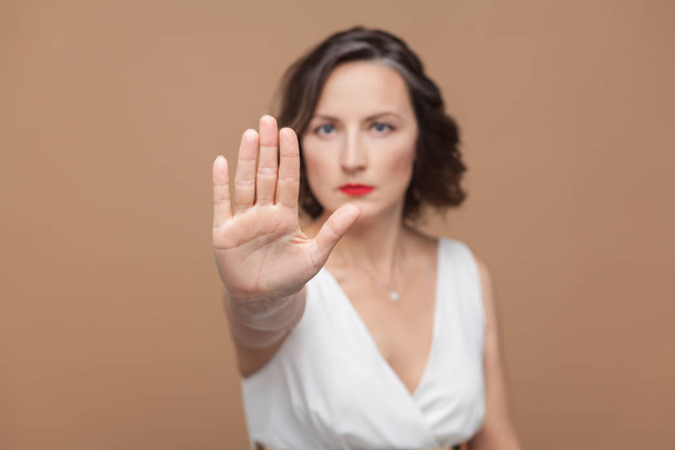 Woman in white dress showing hand ban sign at camera on brown background  - Fotó, kép