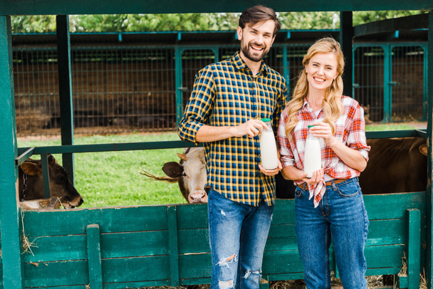 sonrientes parejas de granjeros sosteniendo botellas de leche de vaca cerca de establo
 - Foto, Imagen