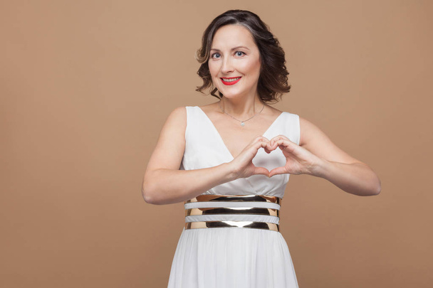 Happy woman in white dress showing heart shape by hands on brown background - Valokuva, kuva